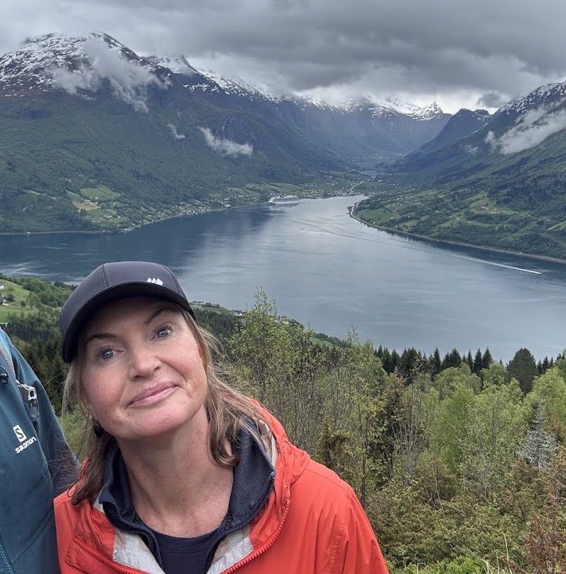 Lena smiling on a mountainside, with a fjord and snow-capped peaks behind her